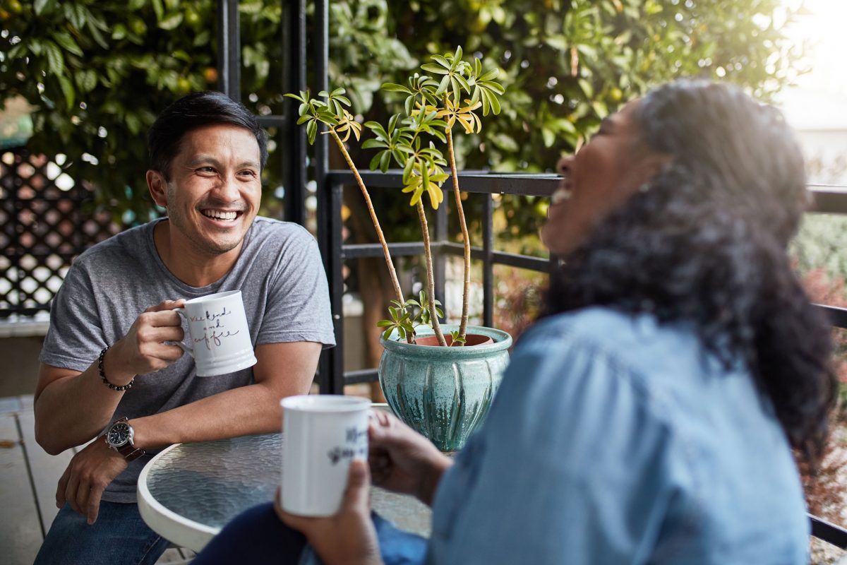 older man and woman drinking coffee and laughing outdoors surrounded by greenery