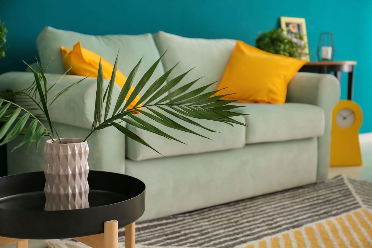 pistachio colored couch with turquoise accent wall, yellow pillows, round side table and beachy greenery